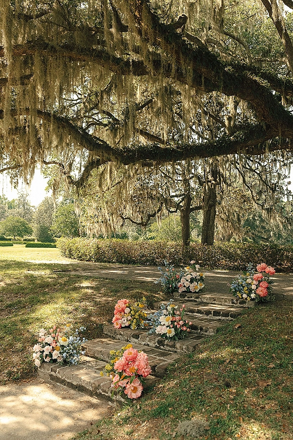 Outdoor ceremony aisle with flower lined aisle markers on stone steps, leading to an oak tree draped in hanging moss in sunlight