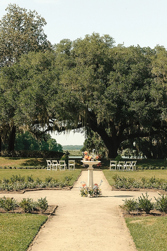 Ceremony setup with white folding chairs lining a garden aisle, pedestal urn florals at the end beneath oak trees and hanging moss
