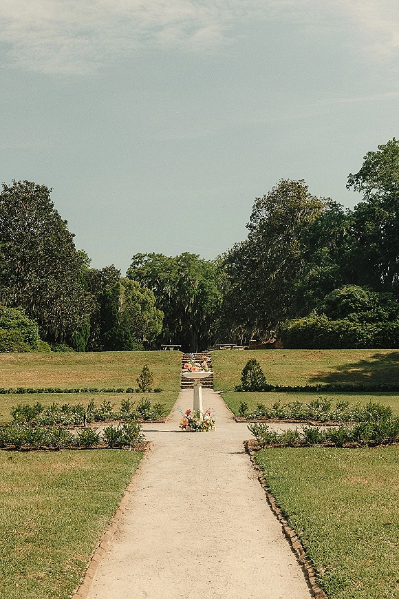 Ceremony aisle decor with wedding aisle flowers in pedestal urns lining stone steps and brick-edged path across a garden lawn with hedges