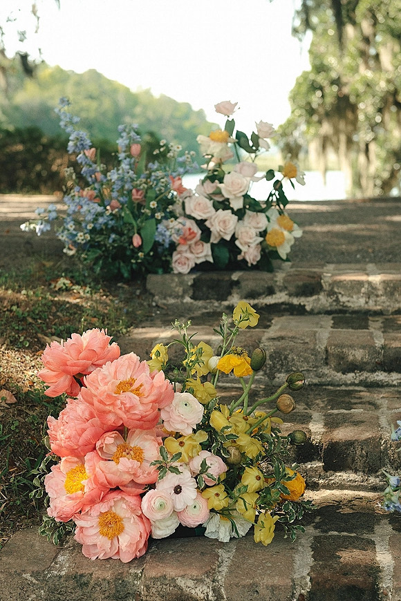 Wedding floral arrangement of coral peonies and blush roses with greenery on stone steps beside a garden lawn and water backdrop