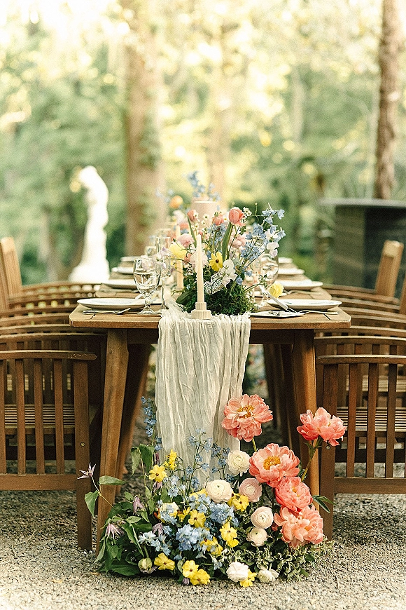 Reception tablescape with an outdoor reception table on a wood dining table, featuring wildflowers, taper candle, and gauze runner in a garden setting