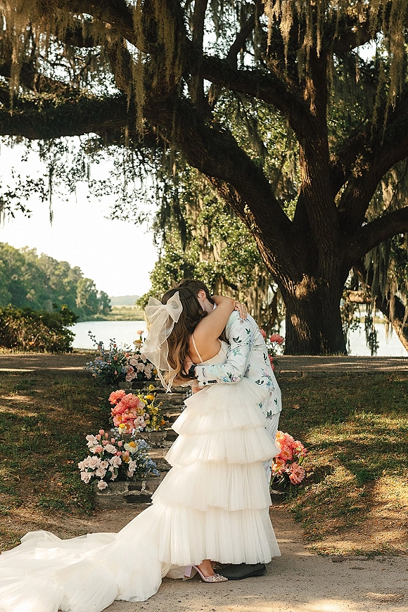 Wedding kiss portrait of bride and groom kissing, her veil bow and tiered dress flowing on stone steps under an oak by lakeside water