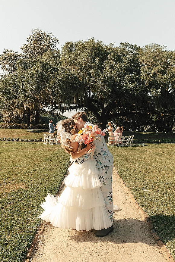 Wedding kiss as bride in a veil and tiered tulle dress kisses groom in floral suit, holding a colorful bouquet on oak-lined lawn aisle