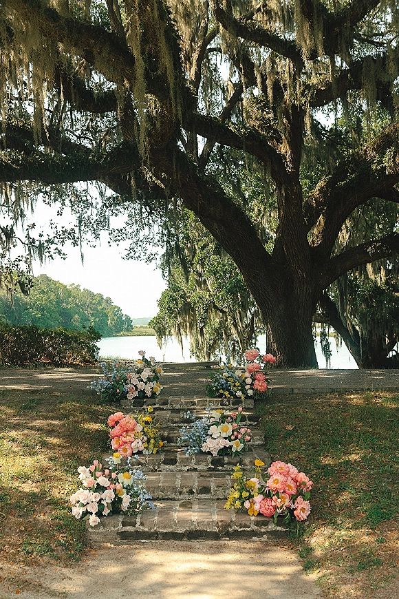 Ceremony aisle decor with outdoor ceremony aisle flowers in ground floral arrangements on stone steps beneath a mossy oak by the lake
