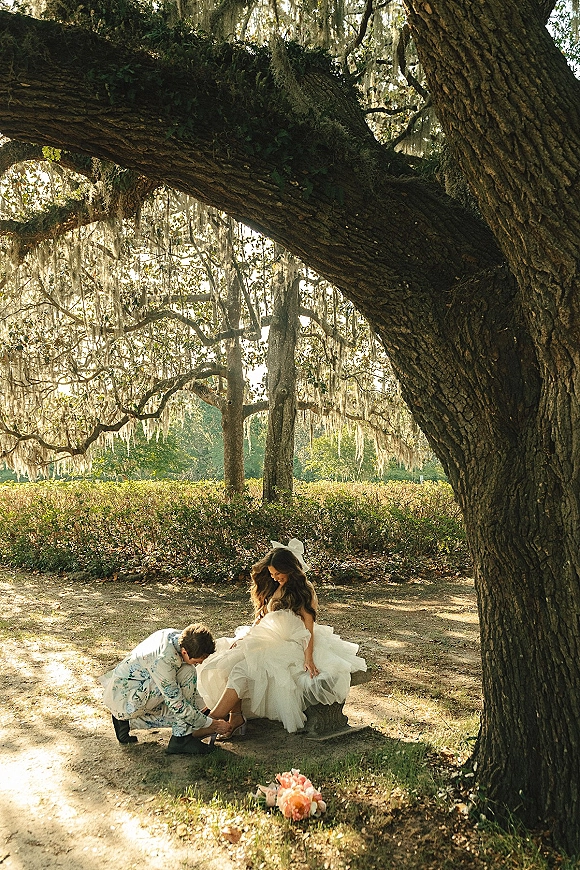 Couple portrait of bride laughing on a stone bench as groom adjusts her heel, with bouquet nearby under sunlit oak trees and moss