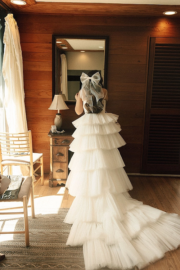 Wedding dress with tiered tulle skirt and long train, back view in a wood-paneled room with mirror, lamp, and window light