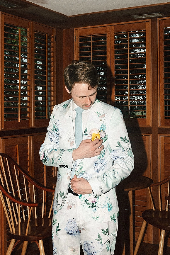 Groom portrait in a floral suit adjusting his boutonniere, wearing a light blue tie in a wood-paneled room by a window