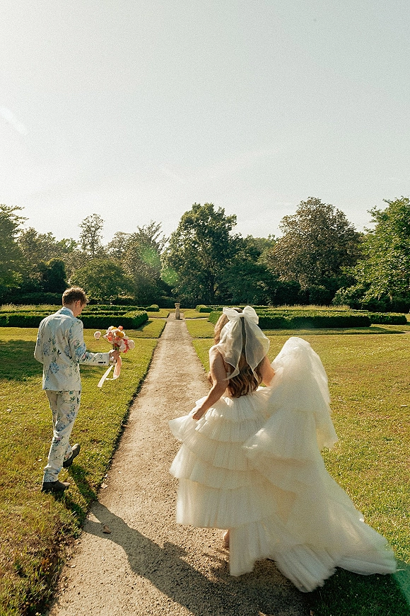Couple walking portrait, bride and groom walking away on a sunlit garden path, her long veil and bouquet with ribbon trailing behind
