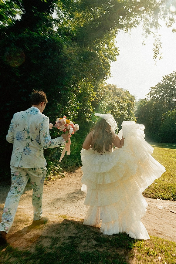 Wedding couple portrait of bride lifting her ruffled dress and groom holding a colorful bouquet on a sunlit garden path, with veil bow