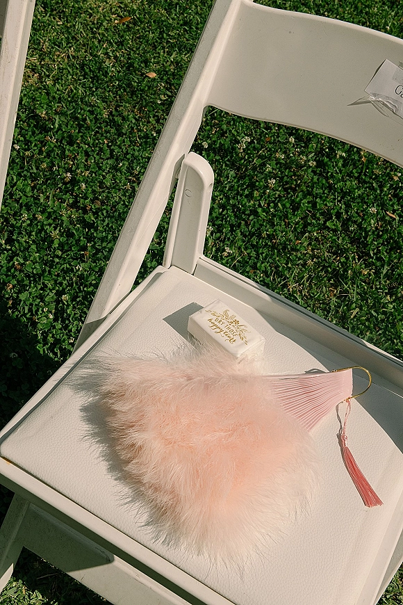 Wedding ceremony favor with wrapped soap, blush feather fan, and tassel set on a white folding chair on a grass lawn outdoors
