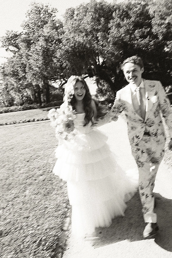 Wedding couple portrait in a black and white wedding photo, bride and groom walking hand in hand on a sunlit garden path with veil
