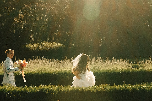 Couple portrait of bride running beside groom holding bouquet, her veil and ribbon streamers flying in sunlit garden meadow grass