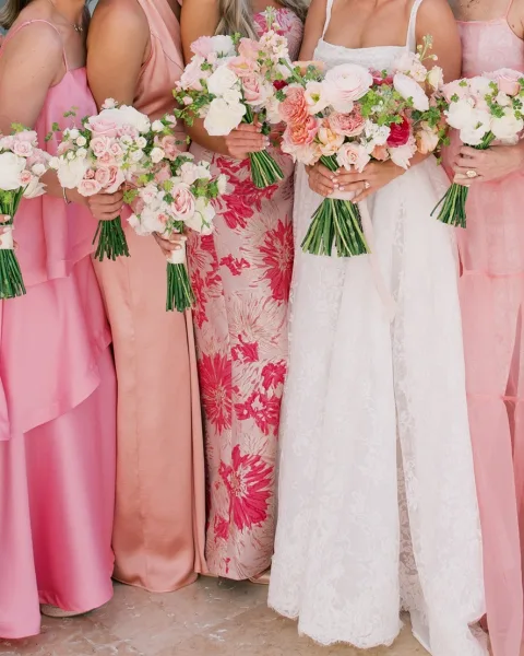 Bridesmaid bouquets of pink and white wedding bouquets with roses, ranunculus, and greenery held beside mix-and-match dresses on a neutral floor