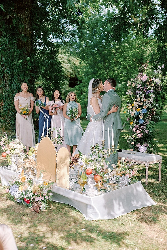 Ceremony kiss as bride in lace gown and veil kisses groom in sage suit beneath floral arch, wedding party behind on garden lawn