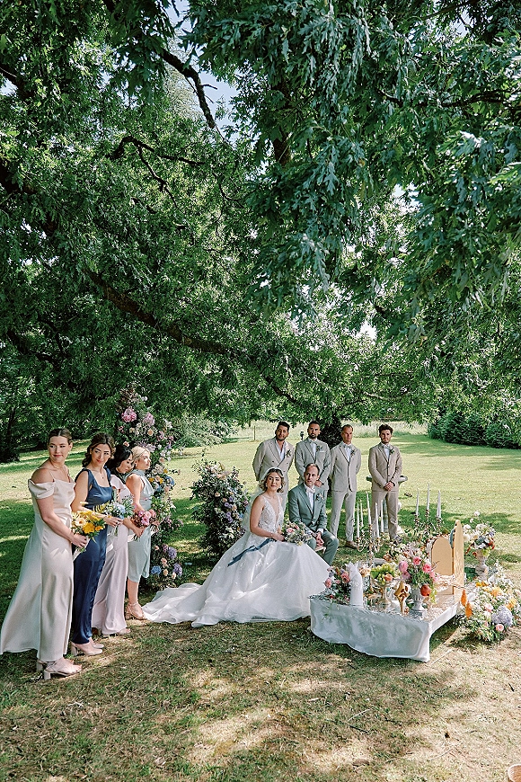Wedding party portrait under a large tree with bride and groom beside bridesmaids in mismatched dresses, picnic table with fruit display and candles
