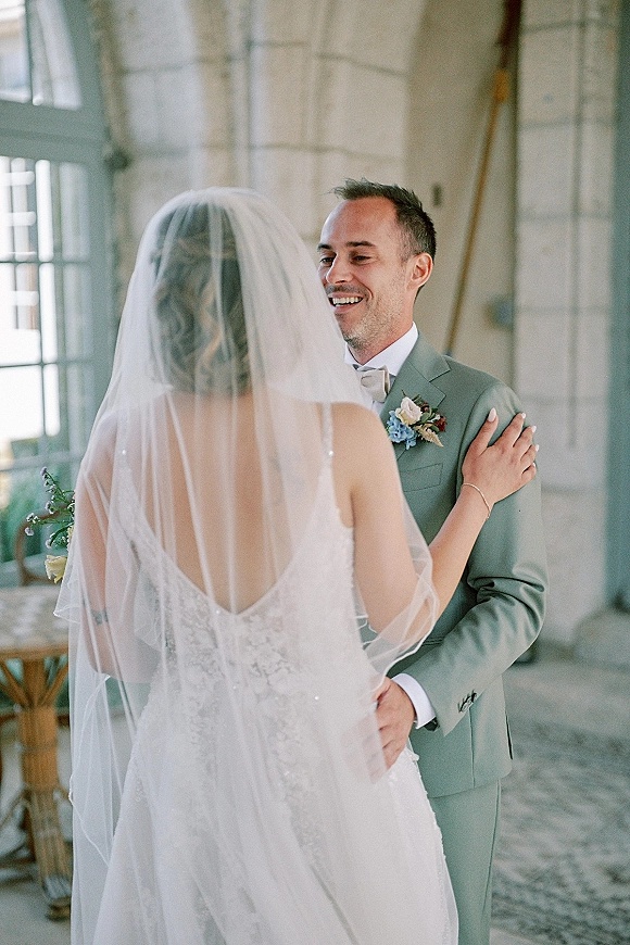 First look moment as bride in lace dress and veil touches groom in bow tie, holding bouquet, in window-lit industrial interior