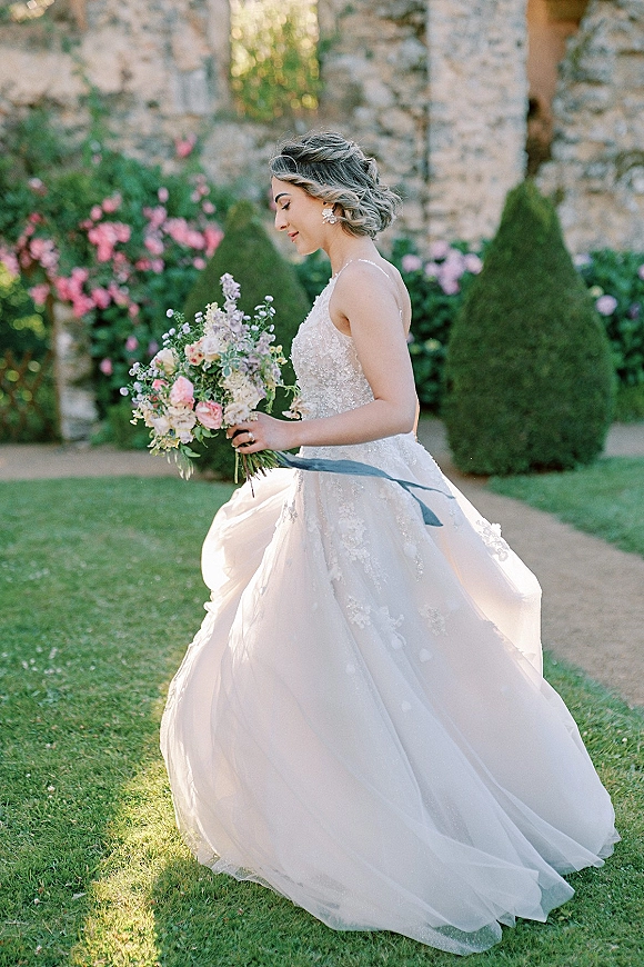 Bridal portrait of bride holding bouquet in side profile, lace bodice and tulle skirt flowing with a blue ribbon on a garden lawn by a stone wall
