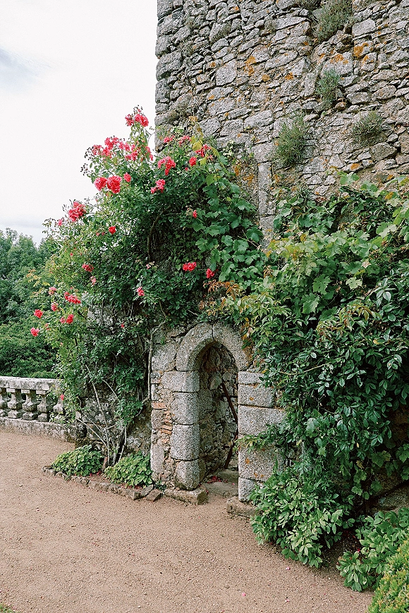 Stone garden archway framed by climbing roses and green vines, with a gravel path leading through the stone arch entrance under trees and sky