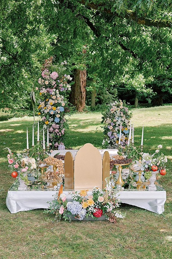 Ceremony backdrop decor with floral installations and taper candles on brass candelabras, set on mirrored platforms with gold panels outdoors