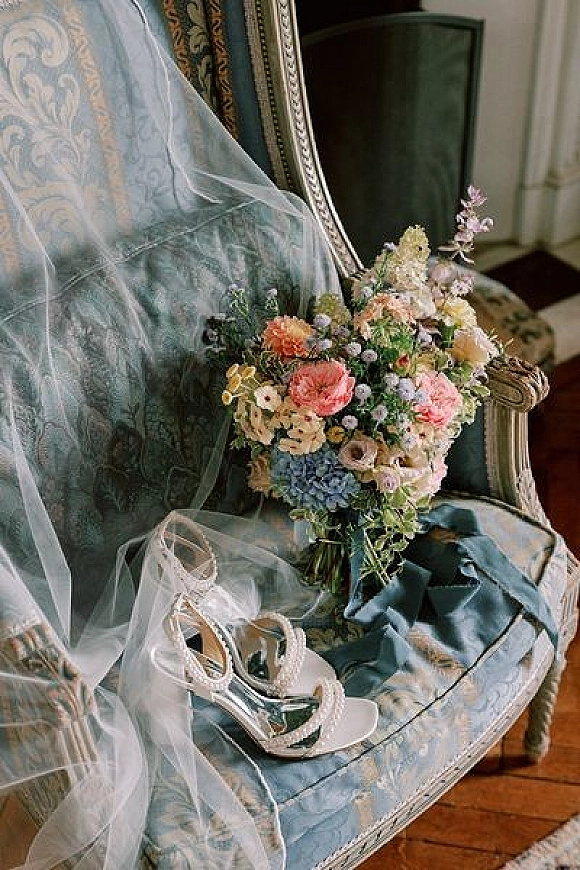 Bridal accessories flat lay with a wedding bouquet and shoes, veil and ribbon arranged on an upholstered chair over a wood floor