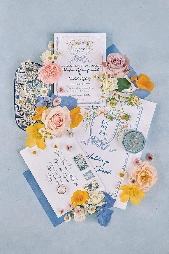 Invitation suite wedding invitation flat lay with blue envelopes, floral illustrations, roses, stamps, wax seal, and ring box on light blue surface