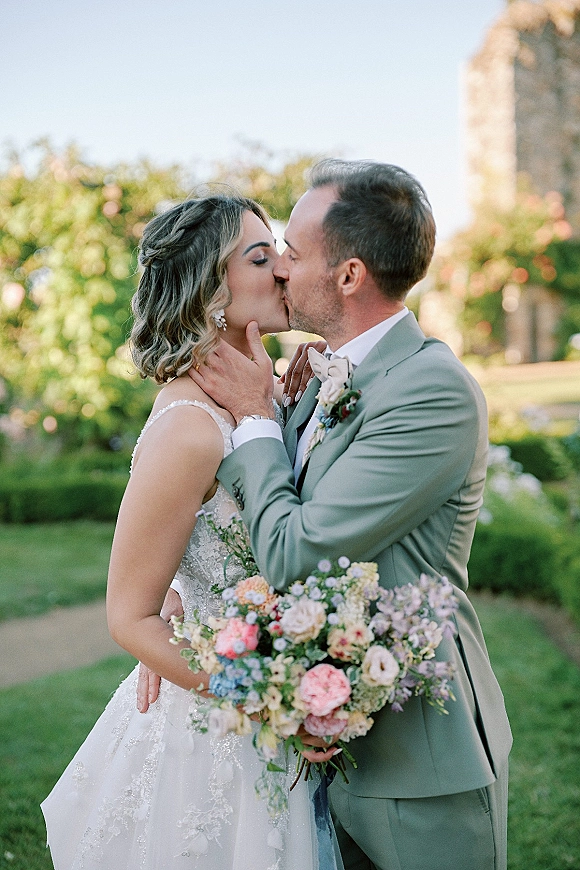 Wedding kiss portrait of bride and groom kissing as he holds her face, bouquet in hand, in a garden with trees and stone building backdrop