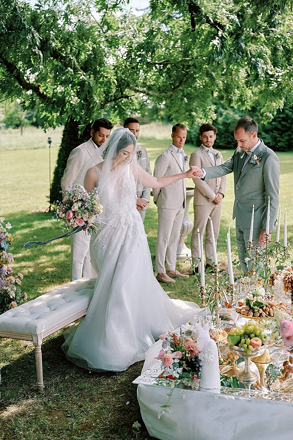 Ceremony moment at an outdoor wedding ceremony as the bride in veil and lace gown walks to the groom on a garden lawn under a large tree