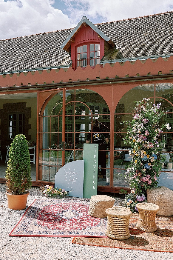 Wedding welcome sign with welcome sign florals beside layered rugs, woven poufs, and a floral pillar at glass-door venue entrance