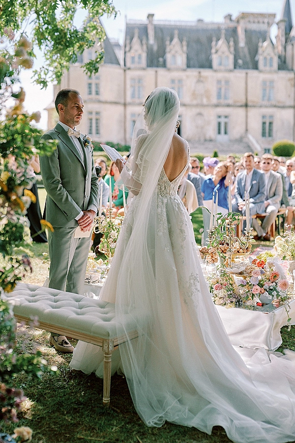 Wedding vows as the bride reading vows from a card beside the groom, lace dress and veil framed by garden guests and manor backdrop
