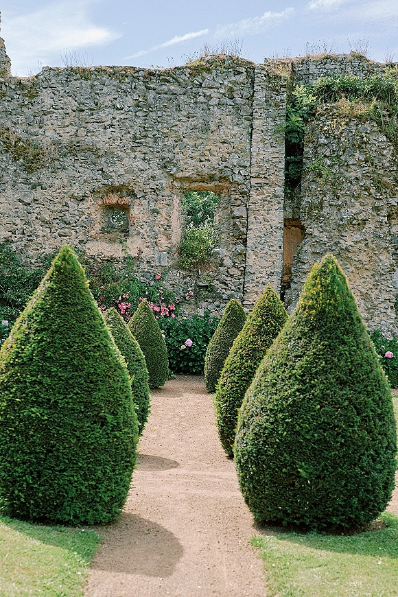 Garden pathway with trimmed topiary shrubs and pink flowers, leading toward stone ruins with an arched window opening under open sky