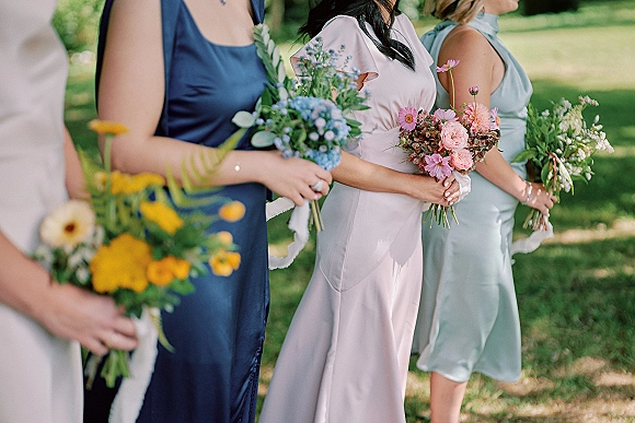 Bridesmaid bouquets in mixed wildflower bridesmaid bouquets style, held by bridesmaids in satin dresses on an outdoor lawn with trees behind