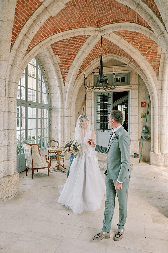 Wedding couple portrait of bride and groom holding hands, her veil and bouquet glowing under a chandelier in a stone-arched hallway.