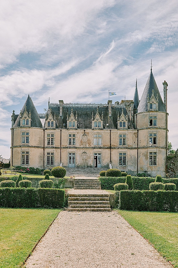 Castle wedding venue with a stone staircase and gravel path, framed by boxwood hedges and ivy, under a blue sky with clouds