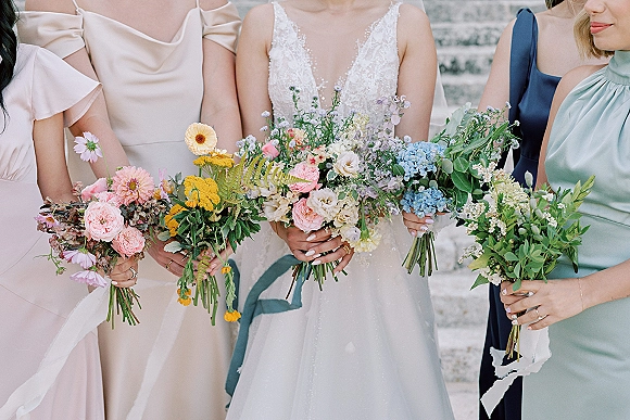Bridesmaid bouquets of wildflower wedding bouquet blooms with greenery and ribbon streamers, held on stone steps beside lace dress and rings