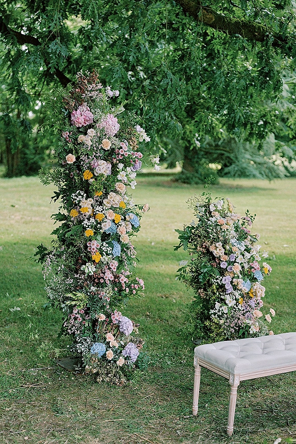 Ceremony floral arch with asymmetrical pastel hydrangeas and roses framing a white tufted bench beneath sprawling garden trees on lawn