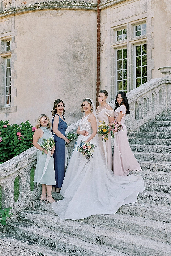 Bridal party portrait of a bride with bridesmaids holding bouquets on a stone staircase outside a historic building with pink flowers