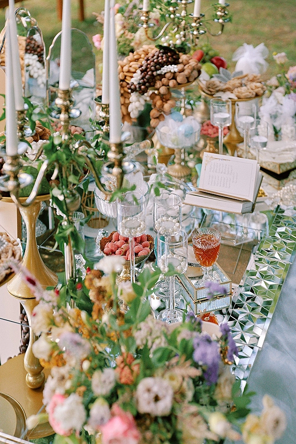 Reception tablescape on a mirrored wedding table with gold candlesticks, white tapers, pastel florals, crystal goblets, and pearl strands on a lawn