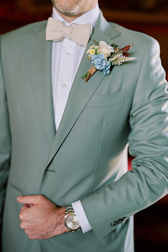 Groom portrait in a sage green groom suit with beige bow tie and boutonniere, wearing a wristwatch in a dark wood interior setting
