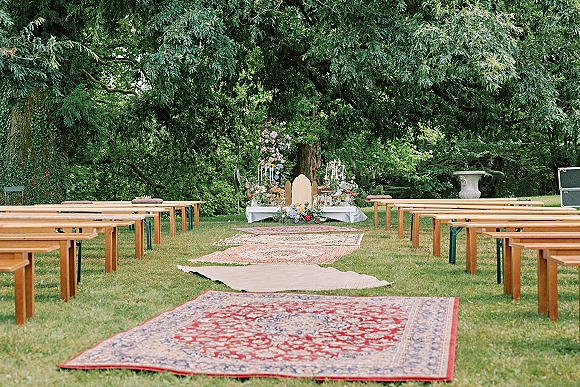 Outdoor ceremony setup with garden wedding ceremony aisle rugs, wood benches, taper candles, and floral altar on a grassy lawn under trees