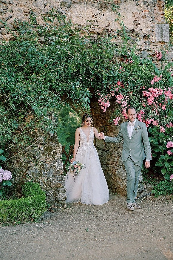Couple portrait of bride and groom walking hand in hand under a stone archway, bride holding a bouquet with greenery and pink flowers