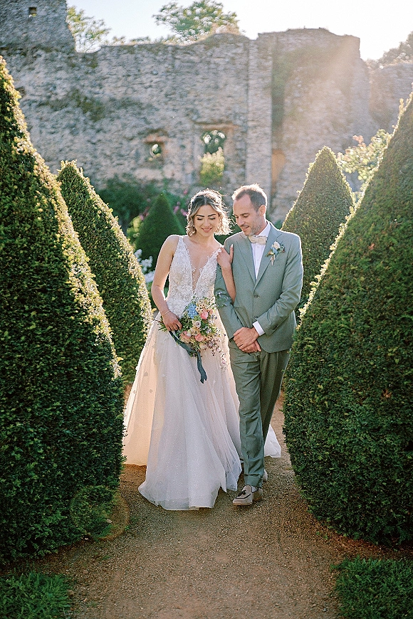 Couple portrait of bride and groom walking on a gravel path in a formal topiary garden, bride holding bouquet, sunlight on stone ruins.