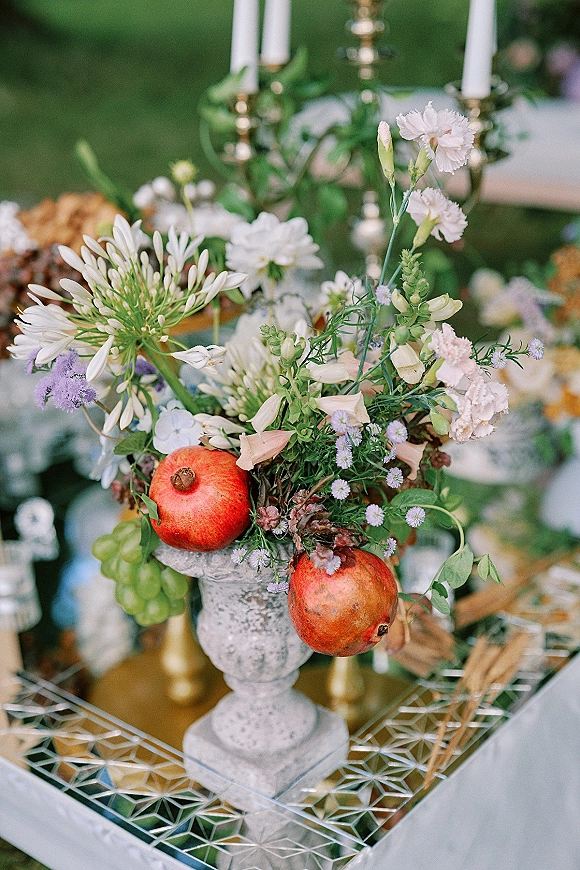Wedding centerpiece with a floral urn arrangement, pomegranates, and gold taper candlesticks on a mirrored tray at an outdoor lawn reception table