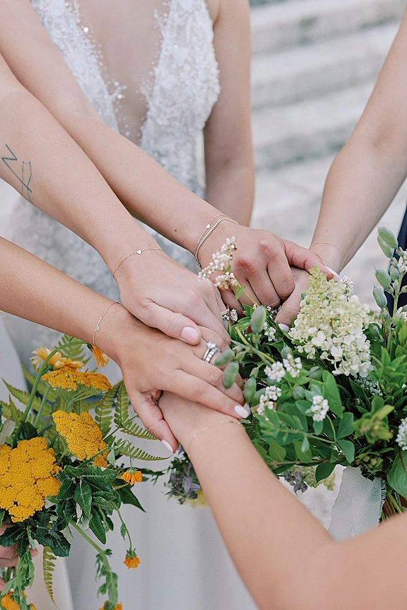 Bridesmaid moment with bridal party hands stack, showing mixed metal wedding rings, gold bracelets, and wildflower bouquets on outdoor steps