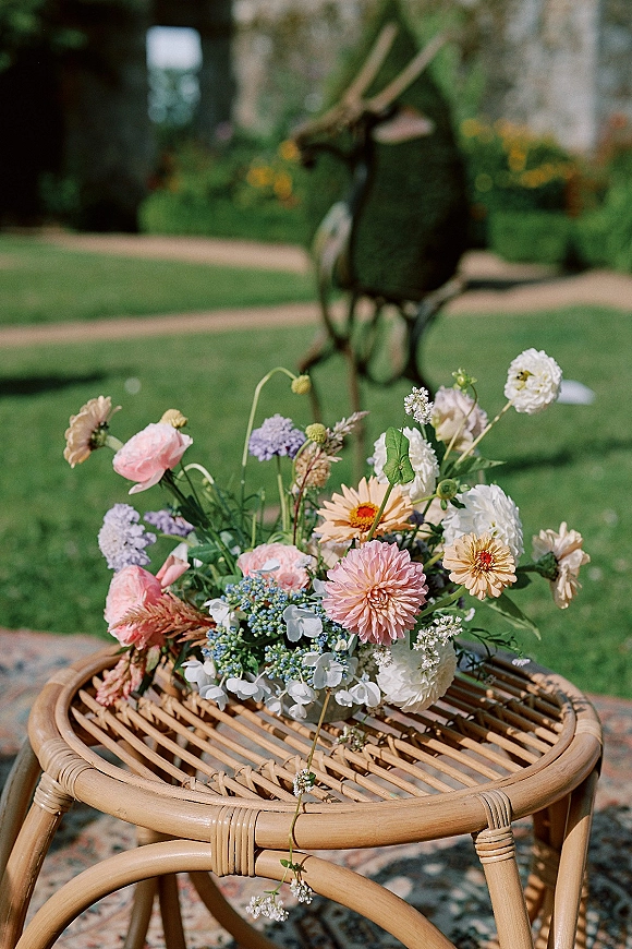 Wedding bouquet of pastel wildflowers with white, pink, peach, lavender, and blue blooms on a rattan table in a garden setting