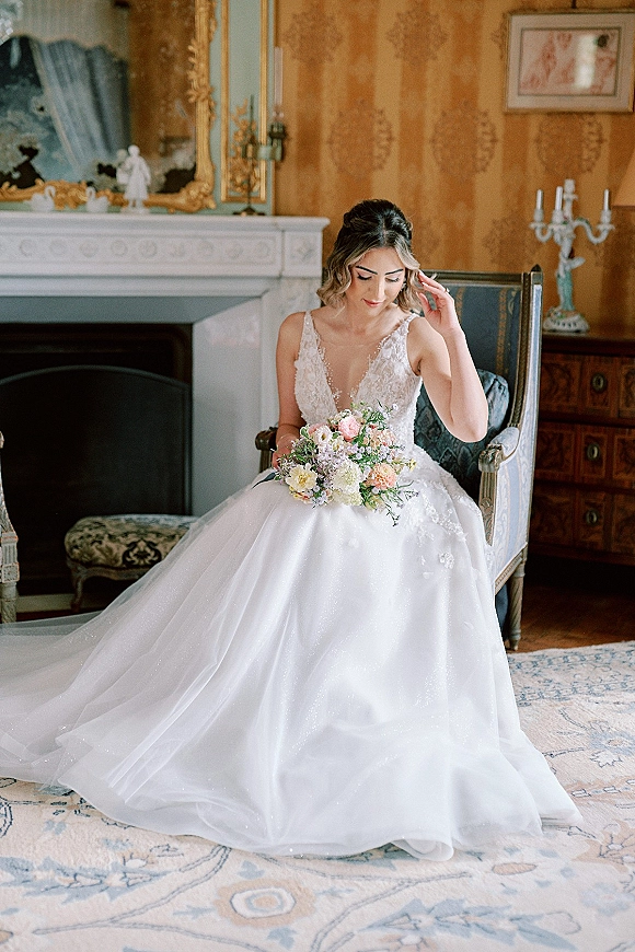 Bridal portrait of a seated bride holding bouquet in a white ball gown with lace bodice, posed by a fireplace and gilded mirror
