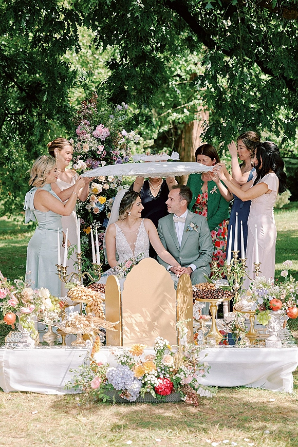Ceremony moment at an outdoor wedding ceremony with bride and groom seated beneath a floral arch, candlelit gold candelabras on a garden lawn
