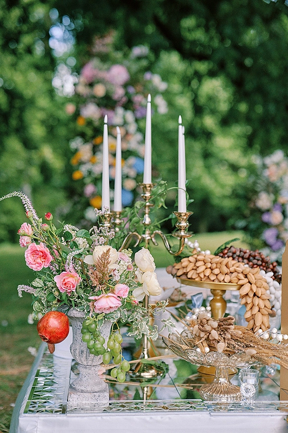Wedding dessert table with roses and greenery, brass candelabra and taper candles on mirrored tabletop, set on a garden lawn near trees