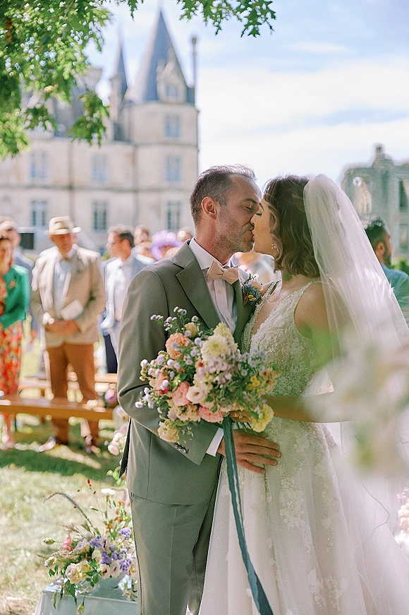 Wedding kiss portrait of bride and groom kissing outdoors, bride holding wildflower bouquet with ribbon, stone castle and guests behind