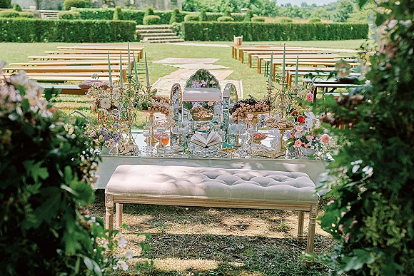 Ceremony altar decor with floral arrangements and taper candles on a mirrored table, set on a garden lawn with hedges and stone steps