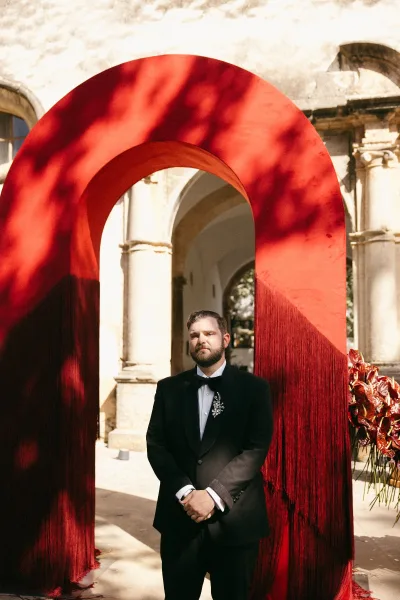 Groom portrait featuring a groom wearing a lapel brooch, posed in a courtyard framed by stone arches and columns.